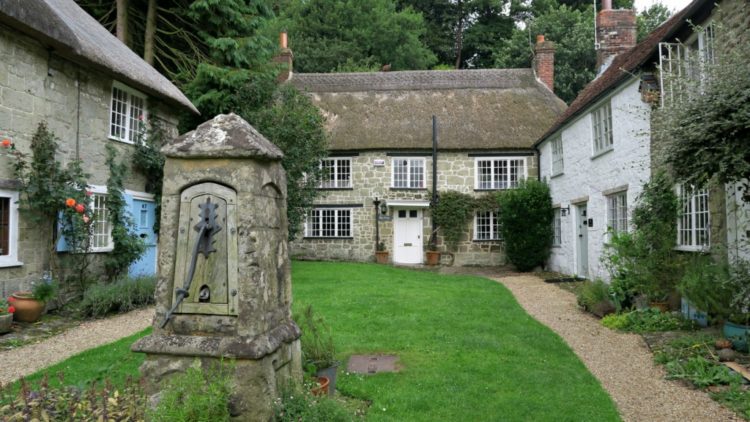 Pump Cottage is a quaint stone cottage with a thatched roof, nestled behind a grassy courtyard. Flanked by flower beds and two other cottages, it features an old stone sundial in the foreground, surrounded by lush greenery and trees.