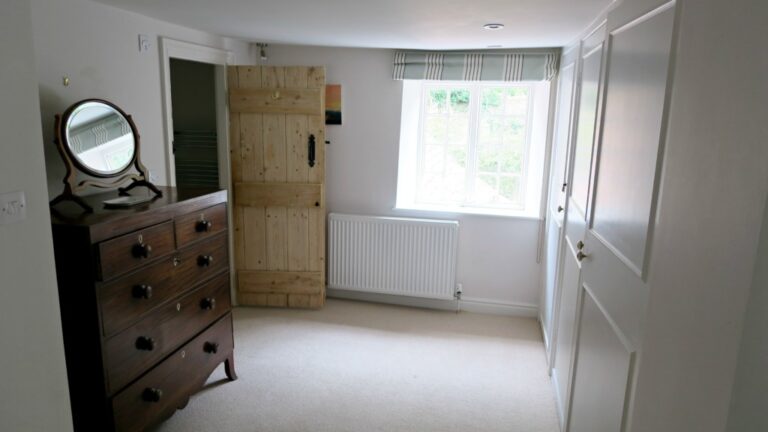 A bright, simple bedroom at Pump Cottage features white walls, a large window, a wooden chest of drawers with a round mirror, a rustic wooden door, and fitted wardrobes. Sunlight fills the space, creating a cosy atmosphere.