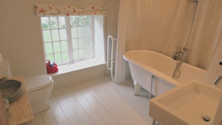 A bright bathroom at Pump Cottage features white walls and floors, a clawfoot bath with shower, a white basin, a toilet, and a window with floral valance letting in natural light. A red bucket sits on the windowsill.