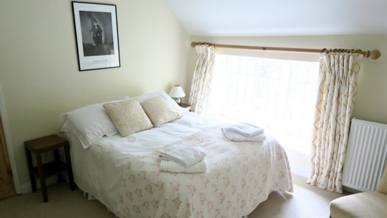 Bright bedroom at Pump Cottage with a double bed, white and pastel bedding, two folded towels on the bed, a small bedside table with a lamp, patterned curtains, and a framed picture on the cream-coloured wall.