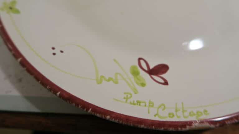 A close-up of a ceramic plate with a red border, green and red leaf-like designs, and the handwritten words "Pump Cottage" near the edge—a charming keepsake from Pump Cottage itself.