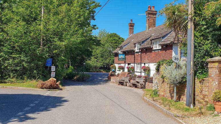 A quiet country road curves past a traditional brick cottage with white windows and hanging flower baskets, surrounded by greenery on a sunny day. Ideal for the Owslebury circular walk, a 30 mph speed limit sign and several roadside signs are visible.