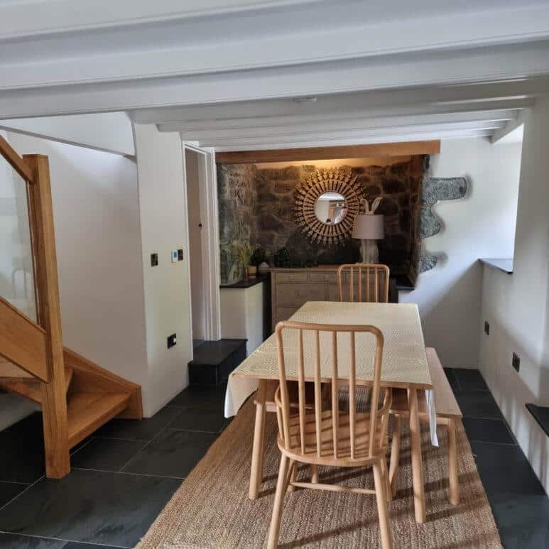 A cosy dining area with a wooden table and four chairs, set on a woven rug. Behind the table is a sideboard with a decorative mirror, lamp, and plants, against a stone feature wall. Wooden stairs are on the left.