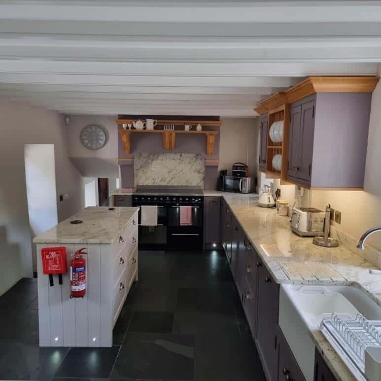 A modern kitchen with marble worktops, purple cupboards, and a central island. There is a cooker with a marble splashback, various kitchen appliances, a sink, and a fire extinguisher mounted on the island.