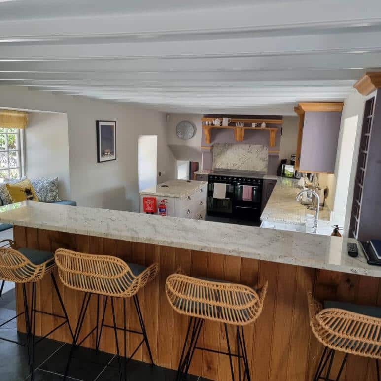 Modern kitchen with a long marble worktop, four wicker bar stools, wooden cabinetry, cooker with overhead extractor, and a cosy seating nook by a window with cushions. Natural light fills the space.