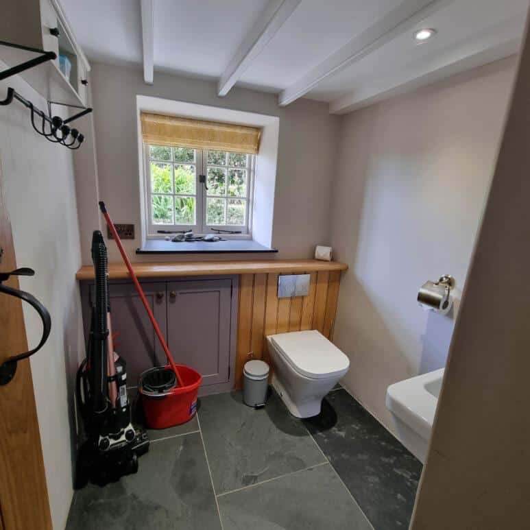 A tidy bathroom with a toilet, wall-mounted flush, small sink, and cupboard under a window. A vacuum cleaner, mop, and red bucket are in the corner. Light streams through the window, illuminating the slate floor.