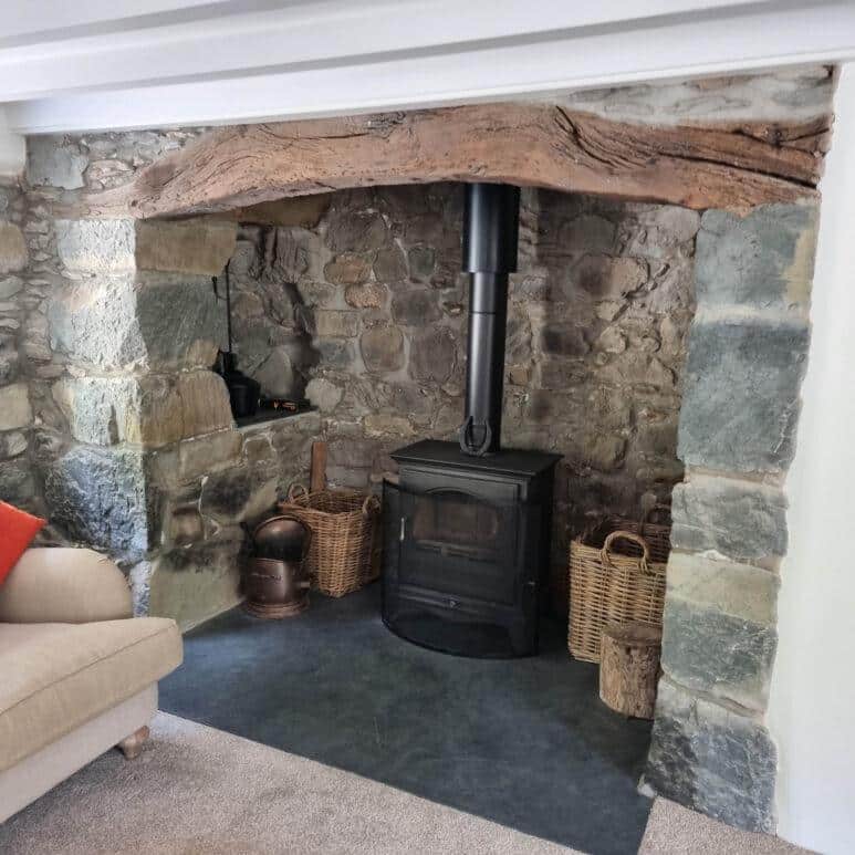 A black wood-burning stove sits in a stone alcove with a rustic wooden beam above. Baskets with logs and kindling are on either side, and part of a beige sofa with an orange cushion is visible on the left.