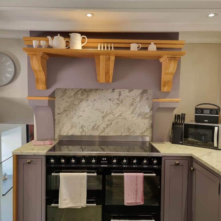 A modern kitchen with a large black cooker, granite splashback, and wooden shelf holding white teapots, cups, and a jug. Tea towels hang from the cooker, and a toaster and utensils sit on the right worktop.