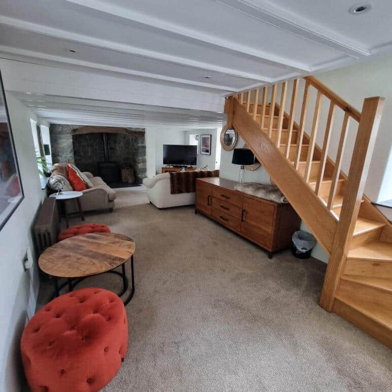 A cosy living room with beige carpet, wooden stairs, modern furniture, two round orange pouffes, a round wooden coffee table, white sofas, and a fireplace with a stone surround in the corner.