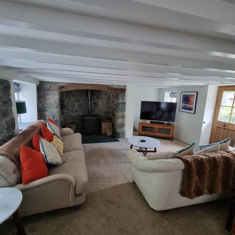 Cosy living room with two sofas, colourful cushions, a stone fireplace with a wood-burning stove, TV on a wooden stand, round coffee tables, and a windowed door letting in natural light.