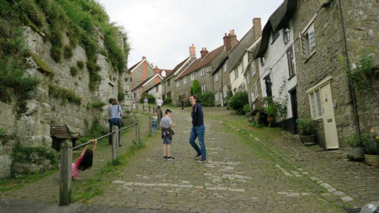 People walk up and down the steep, cobbled Gold Hill, lined with old stone houses on one side and a rocky, grassy wall on the other. One child plays near a railing whilst others make their way up the hill.