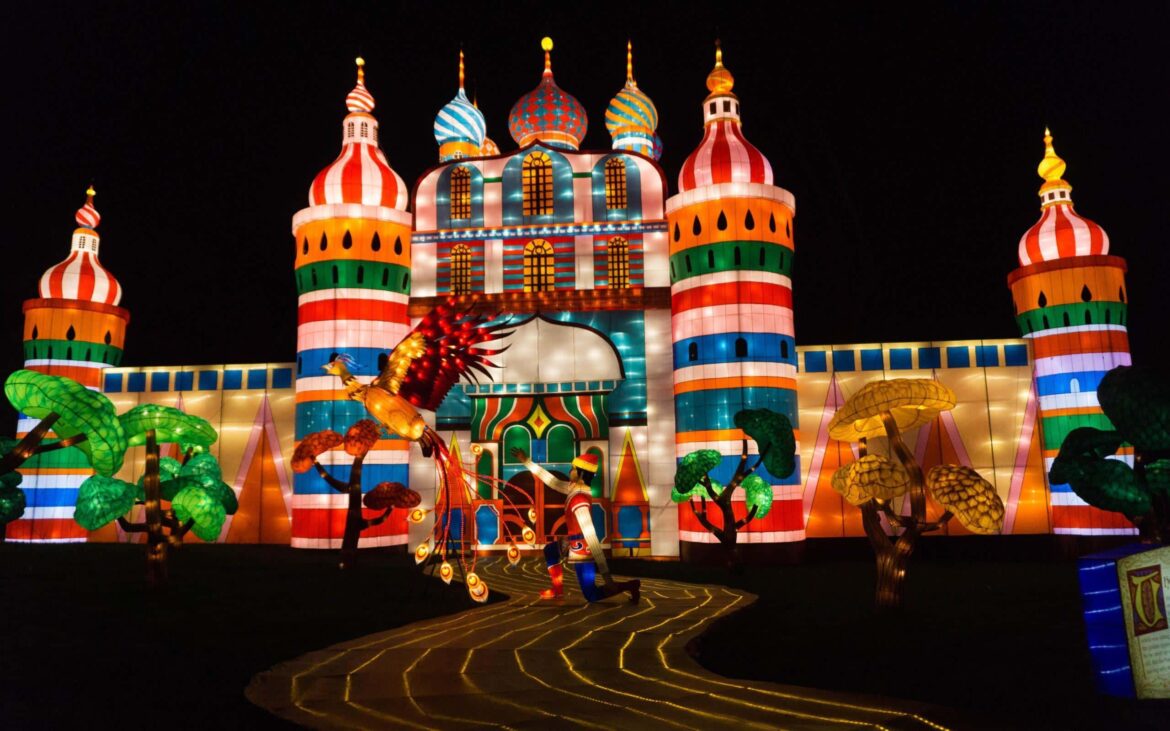 Longleat Festival Of Light and Santa Train A brightly lit, colourful structure resembling a fairytale castle with onion-shaped domes and striped towers, surrounded by illuminated trees and a glowing, fantastical bird on a winding lighted path at night.