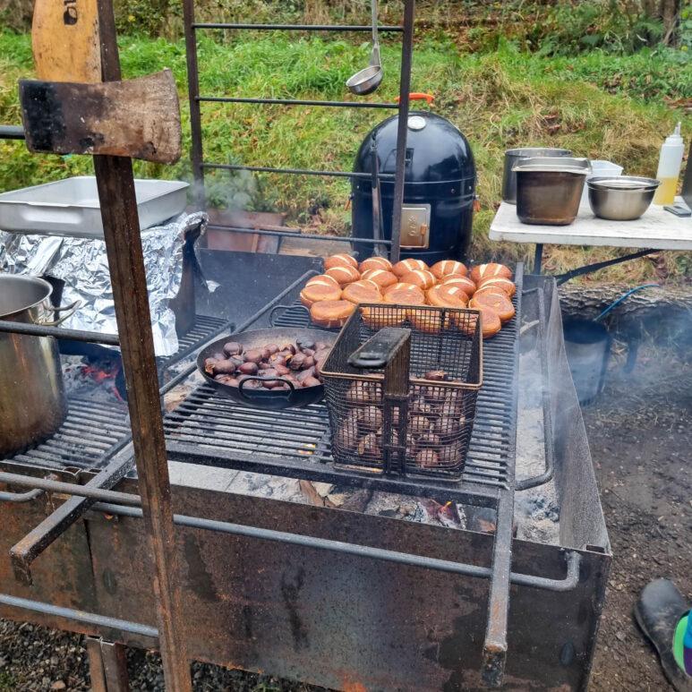 A barbecue outdoors with sliced burger buns, mushrooms in a pan, and a basket of chestnuts cooking. Nearby are pots, bowls, a smoker, and cooking supplies on a table. Smoke rises as the food cooks.