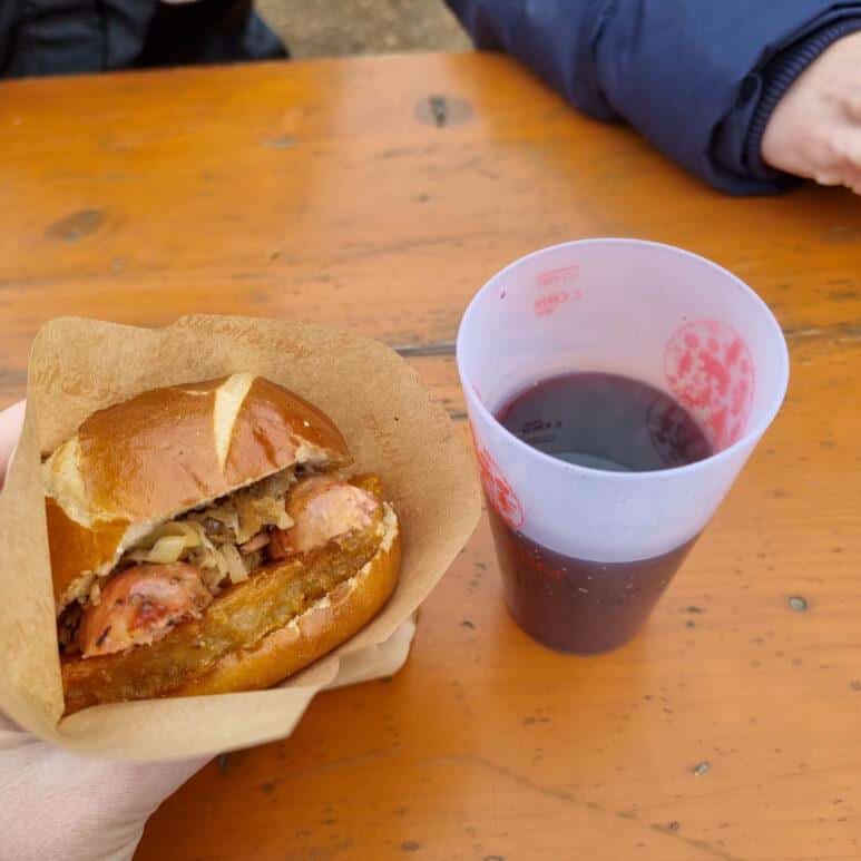 A hand holds a sausage sandwich with sauerkraut in a bap, next to a reusable cup filled with a dark drink on an orange wooden table. Another person's arm rests on the table in the background.