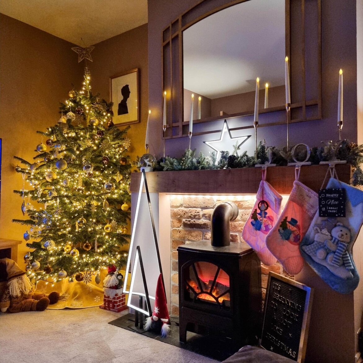 A cosy living room with a decorated Christmas tree, stockings hanging from a mantelpiece above a fireplace, candles, and soft warm lighting creating a festive holiday atmosphere.