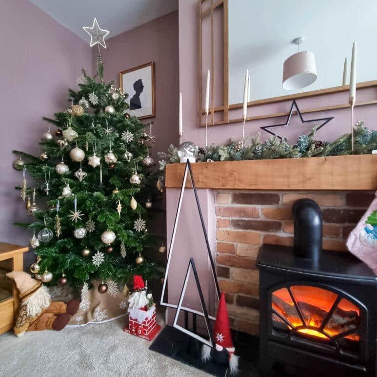 A decorated Christmas tree with gold baubles stands beside a brick fireplace with a lit wood-burning stove. The mantelpiece is adorned with candles, garland, and a star, creating a cosy festive atmosphere.