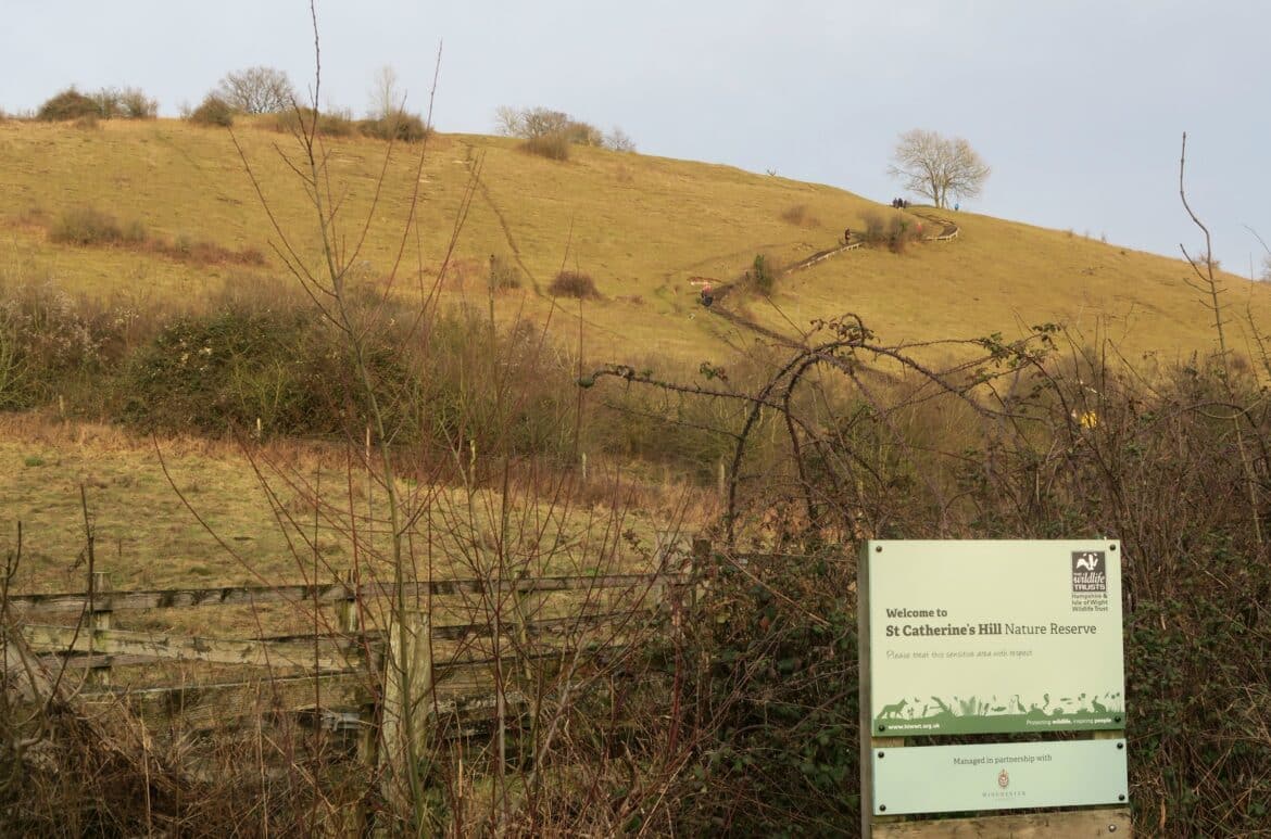 St Catherine’s Hill Winchester A grassy hillside with a winding path leading upward, scattered bushes, and a few trees. In the foreground, a sign reads Welcome to St Catherine’s Hill Nature Reserve.