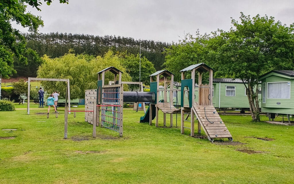 A playground with climbing frames, a slide, and swings on green grass sits within one of the pet-friendly motorhome parks. Two children and an adult stand near the swings. Trees and caravans are in the background on an overcast day.