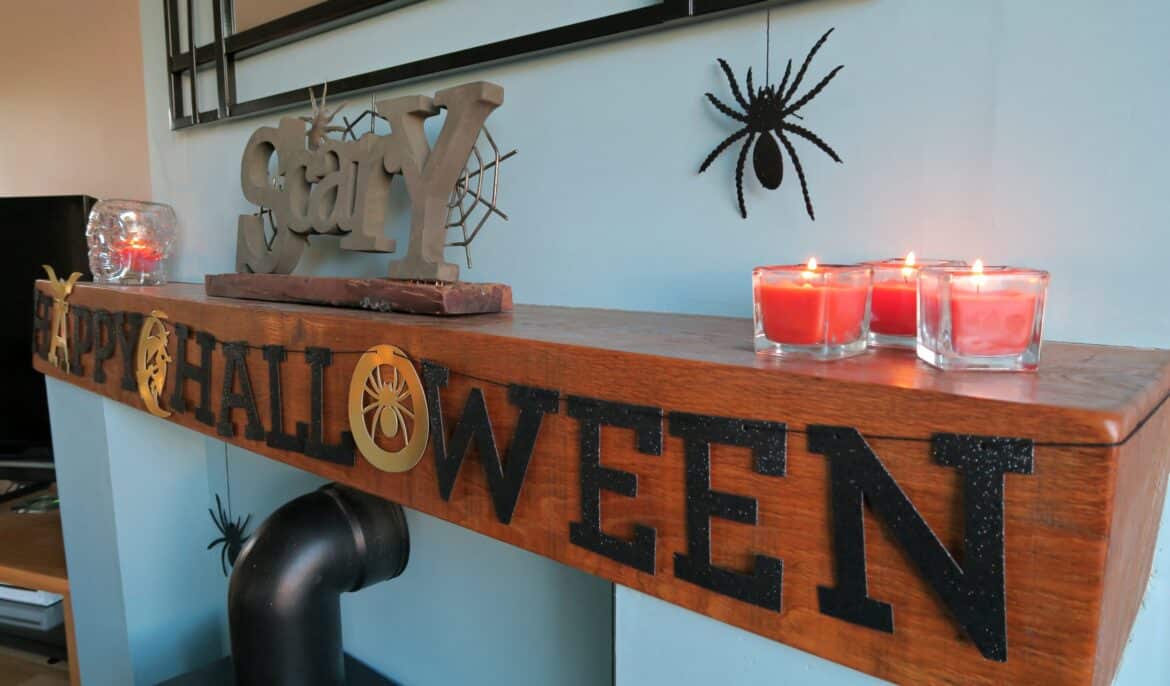 A wooden mantel decorated with black letters spelling “Happy Halloween”, red candles in glass holders, a metallic “stay” sign, glass pumpkin, and black spider decorations creates one of the best destinations for a Halloween gathering.