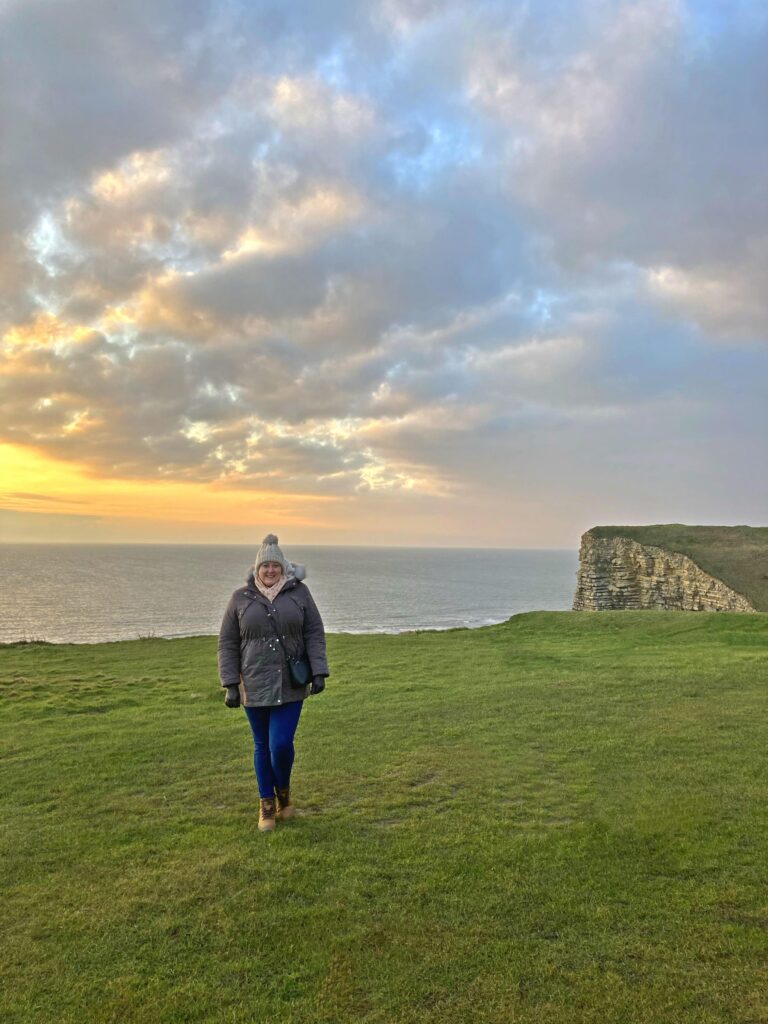 A person in a grey coat, blue jeans, winter hat, and boots stands on a grassy cliff overlooking the sea at sunset, with dramatic clouds and rocky cliffs in the background.