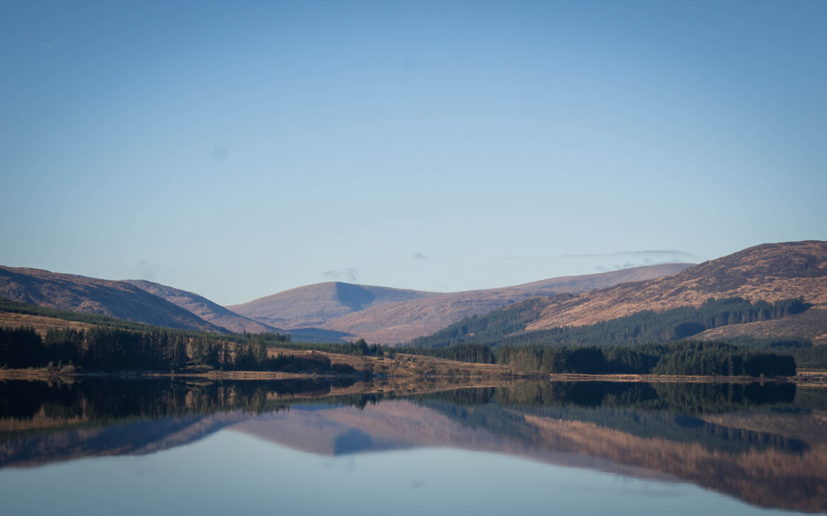 Clatteringshaws Loch in Galloway Forest Scotland A calm lake reflects rolling hills and a line of trees under a clear blue sky, creating a peaceful and symmetrical landscape scene.