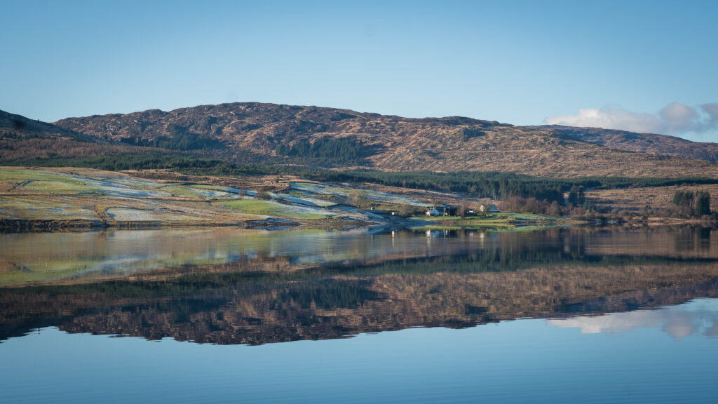 A tranquil lake reflects a hillside with green and brown patches, a few scattered houses, and a clear blue sky above. The scene is peaceful and calm, emphasising nature and open space.