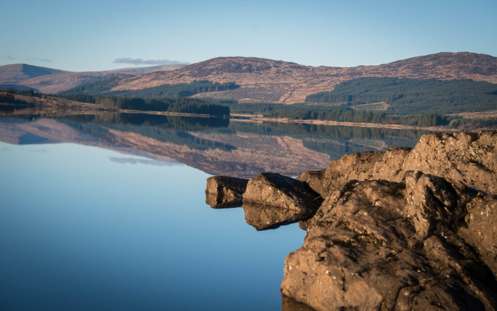 Large rocks sit at the edge of a calm lake, reflecting distant hills and a clear blue sky. Forested slopes and gentle hills are mirrored perfectly on the water’s surface.