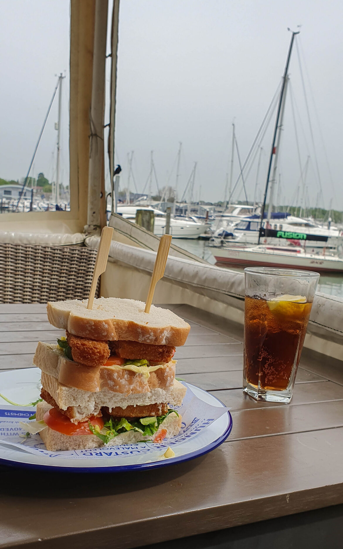 A thick stacked sandwich with lettuce and tomato sits on a plate next to a glass of cola with lemon on a wooden table. Sailing boats and masts, perhaps from local yacht charters, are visible in the marina outside the window.