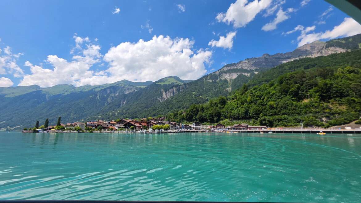 A serene turquoise lake with gentle ripples, bordered by a quaint village and lush green mountains, invites those collecting stories under a bright blue sky with scattered clouds.