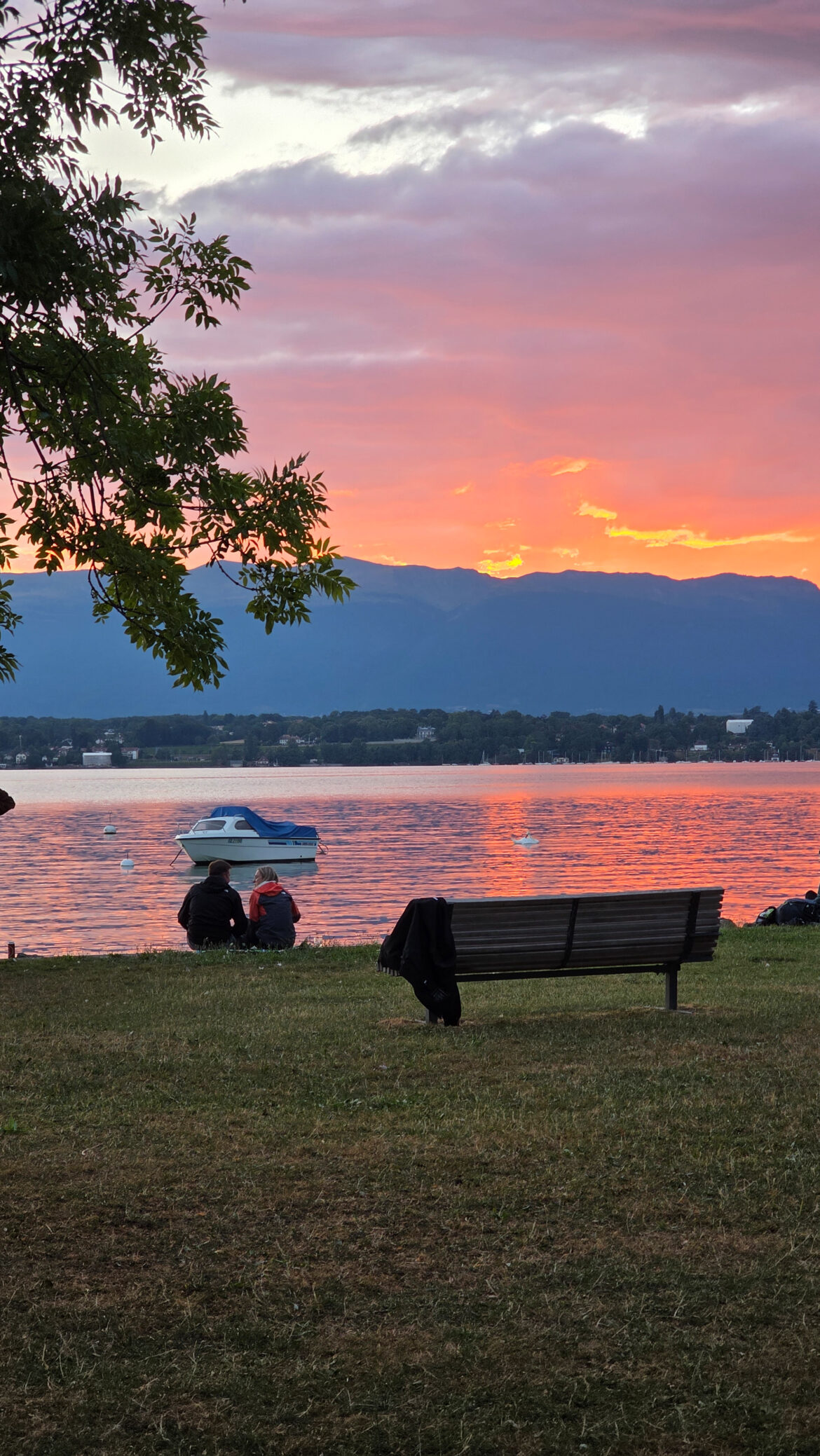 A lakeside scene at sunset with a vibrant orange and pink sky reflecting on the water, a bench in the foreground, and two people—perhaps proposing abroad—sitting near the shore as a small boat floats nearby. Mountains rise in the background.