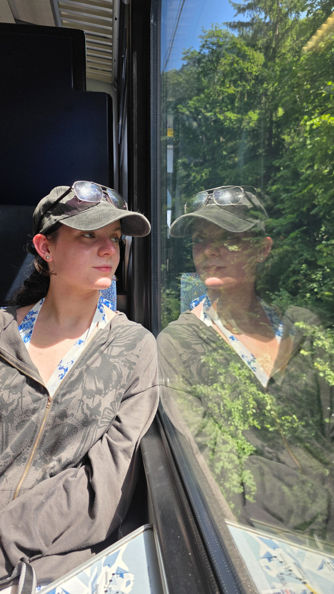 A woman wearing a black cap and sunglasses sits by a train window in the UK, looking at her reflection. Sunlight and green trees are visible outside, creating a bright, serene atmosphere.
