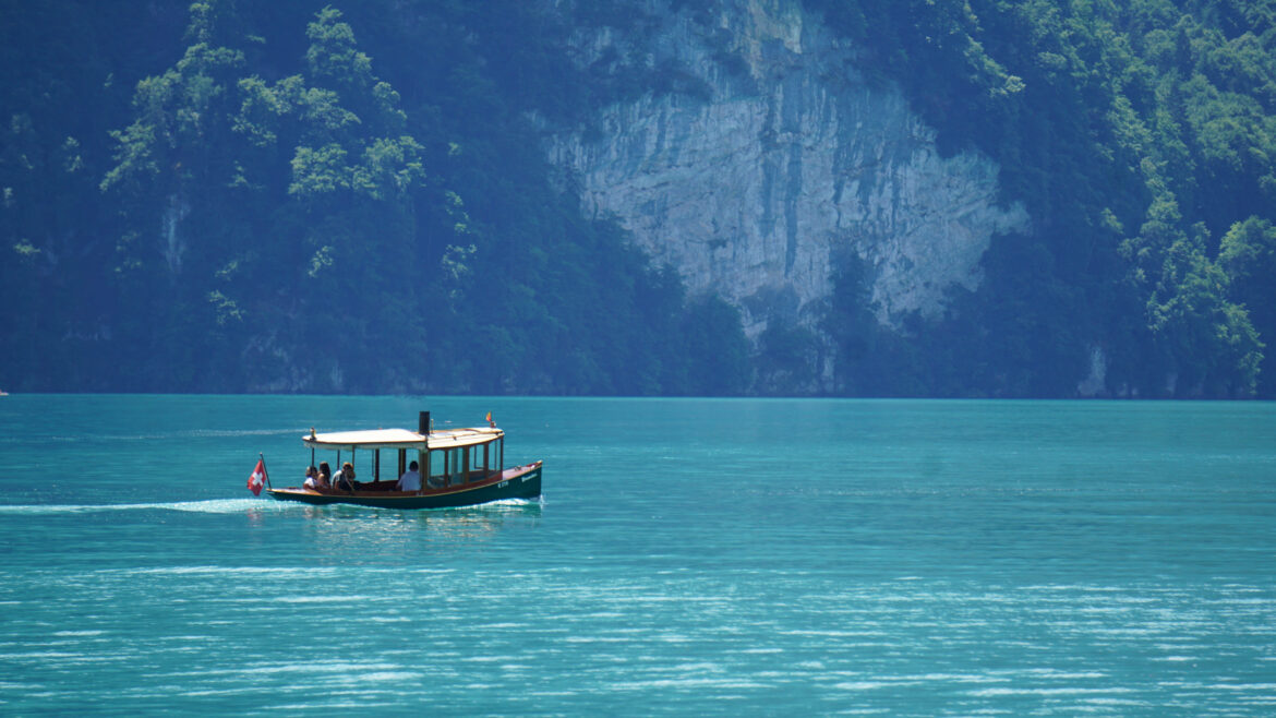 A small boat with a canopy, captained by a confident sailor, sails on a calm, turquoise lake surrounded by lush green mountains and rocky cliffs under bright daylight.