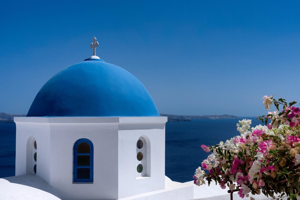 A white building with a blue domed roof and a cross overlooks the blue sea, colourful pink and white flowers in the foreground—capturing a Perfect Day in Santorini with distant islands under a clear sky.