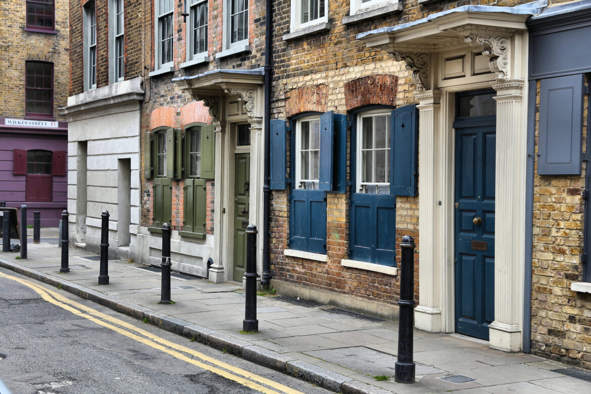 A row of historic brick terraced houses in Shoreditch with colourful doors and shutters, sash windows, and decorative columns lines a quiet, narrow street marked by bollards and double yellow lines.