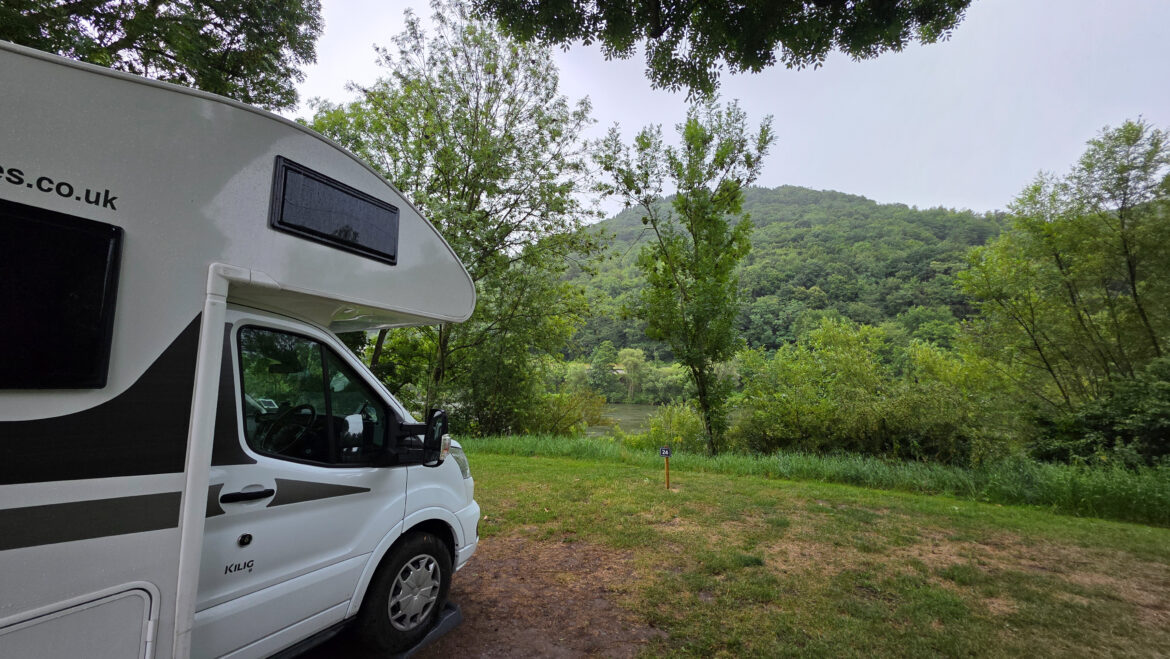 A white camper van, perfect for a family motorhome road trip in the UK, is parked on grass near a river, with trees and a green, forested hill in the background under a cloudy sky.